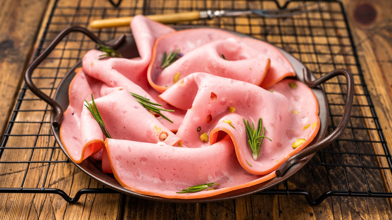 Slices of mortadella with pistachios in a brown bowl with side handles on a black wire rack on a wooden table
