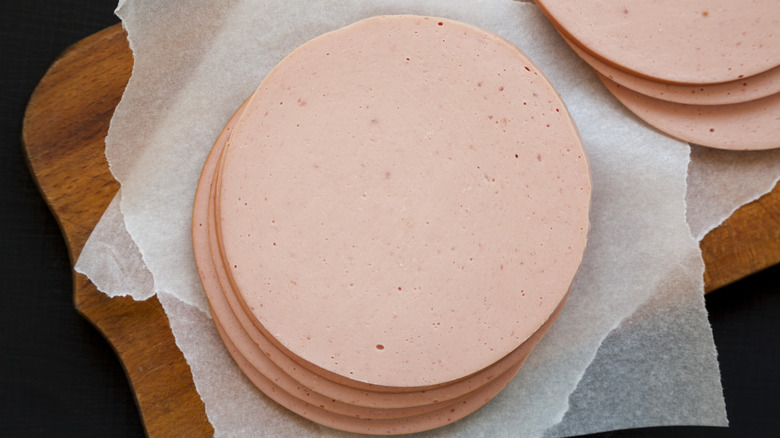 One pile of several round bologna slices with a cut-off image of another pile next to it, both on wax paper on a wooden board