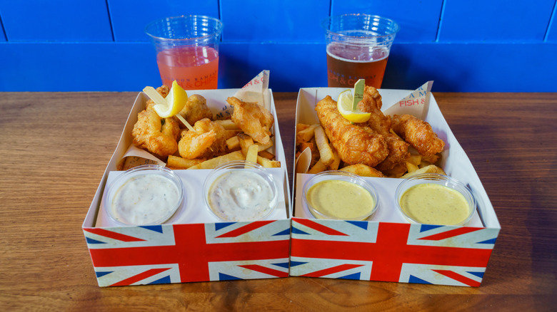 Two orders of fish and chips from Gordon Ramsay's restaurant with drinks and dipping sauces on a wooden table