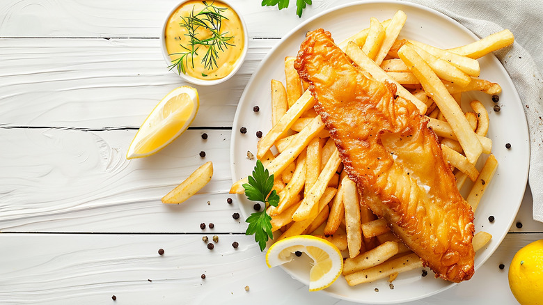 An overhead shot of a white plate of fried fish and fries. with lemon wedges and a cup of sauce on a white table