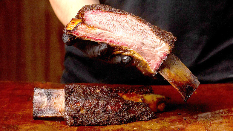 A man holding out beef rib with another on a cutting board.