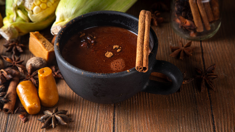 A mug of champurrado on a table with two cinnamon sticks and ears of corn in the background