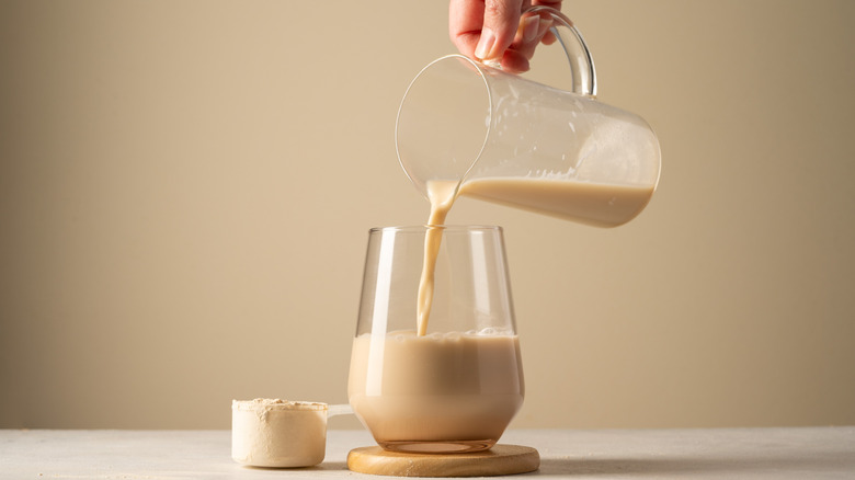 Someone pouring coffee from a glass jug into a glass sitting on a wooden coaster next to a scoop of protein powder
