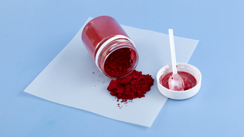 Red powdered dye spilling out of a jar onto a square of white paper with a plastic spoon resting inside the lid nearby