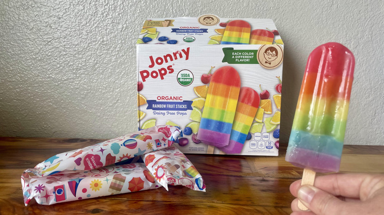 Hand holding a Jonny Pops Organic Rainbow Fruit Stack Popsicle in front of a box of them