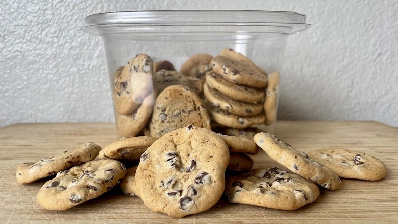 A container of Kirkland Signature Mini Chocolate Chip Cookies with some scattered around it on a wooden cutting board