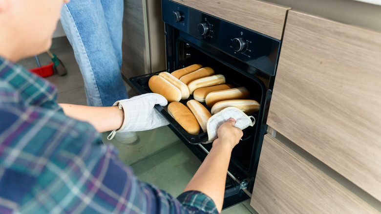 Hot dog buns being pulled out of the oven by a man in a plaid shirt.