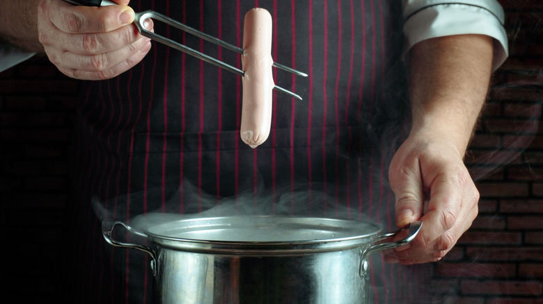 An uncooked hot dog rests on a fork above steam in a pot.