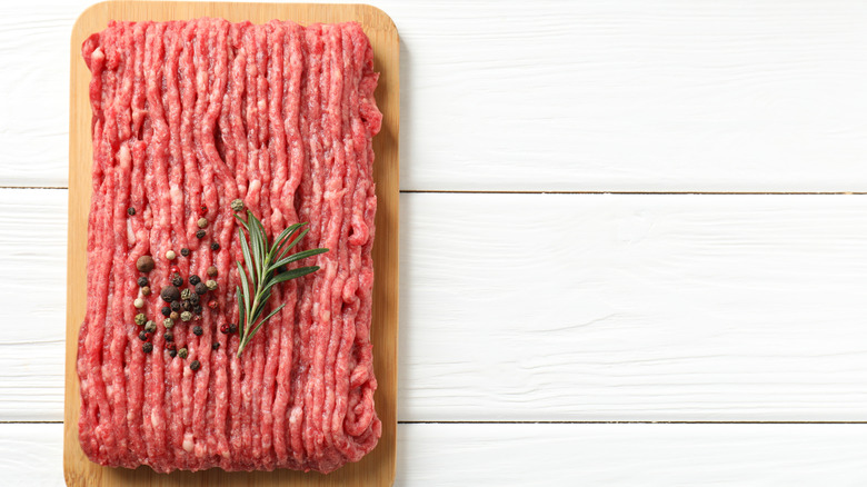 Raw ground meat topped with a sprig of rosemary and some peppercorns on white wooden table, top view.
