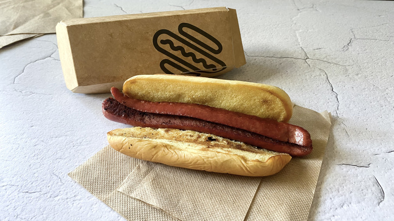 A Shake Shack hot dog on a napkin next to the cardboard takeout container