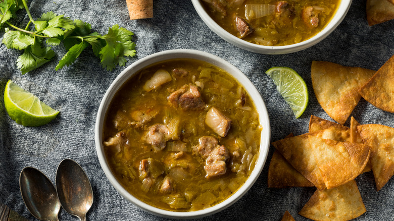 Bowls of Colorado pork green chili stew with tortilla chips on a blue table cloth next to limb wedges, cilantro, and two silver spoons
