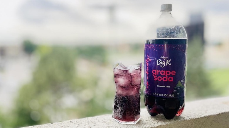 Glass and two liter bottle of Big K Grape Soda sitting on an outdoor ledge