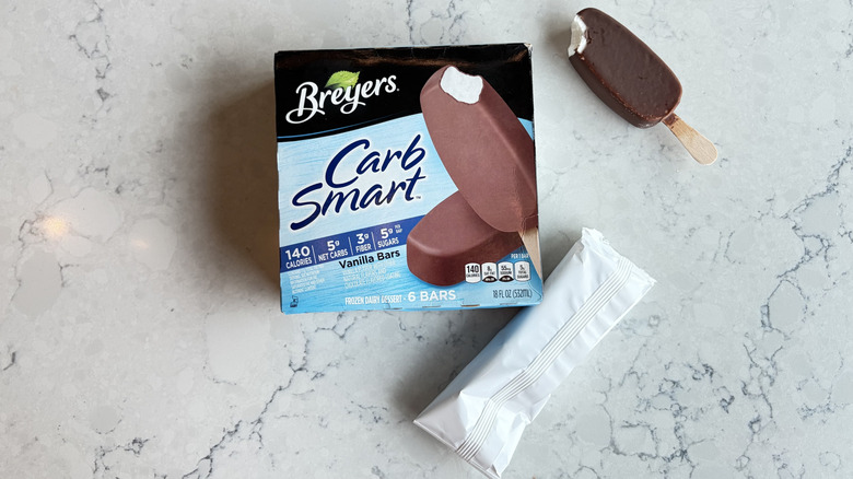 A Breyers Carb Smart Vanilla Frozen Dairy Dessert Bar with a bite taken out of it sits next to its package