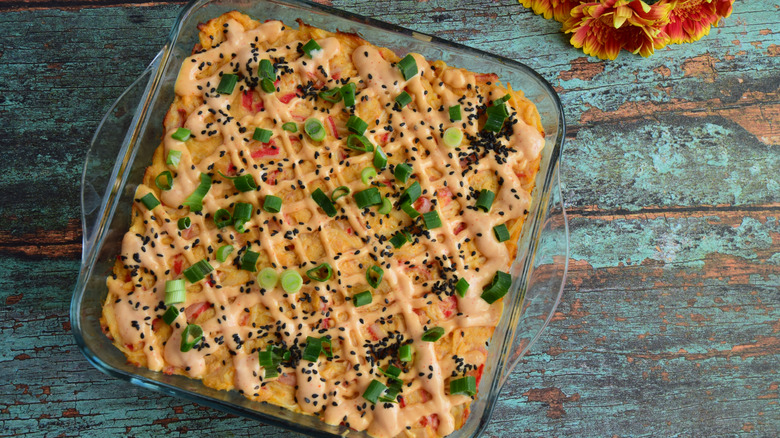 Sushi bake in a glass baking dish, covered with streaks of sriracha mayo, black sesame seeds, and green onions