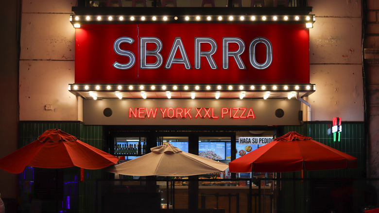 An illuminated Sbarro restaurant sign at night with red and white umbrellas outside the restaurant