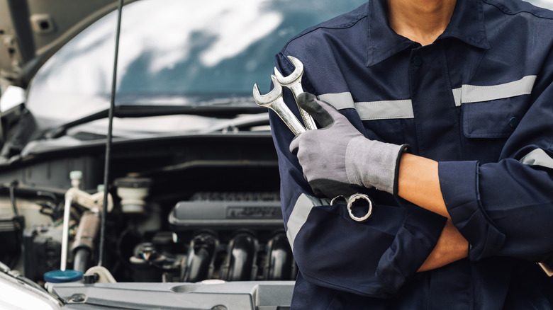An automotive technician holding two wrenches in front a vehicle with the hood popped open.