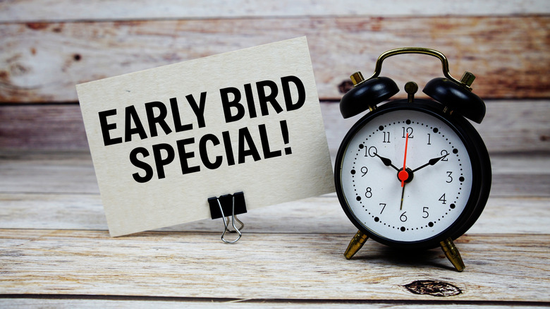An alarm clock with a note next to it that reads "Early Bird Special!" on a wood table.