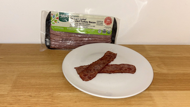 Two pieces of 365 Organic turkey bacon laid across each other on a white plate with the package behind it.