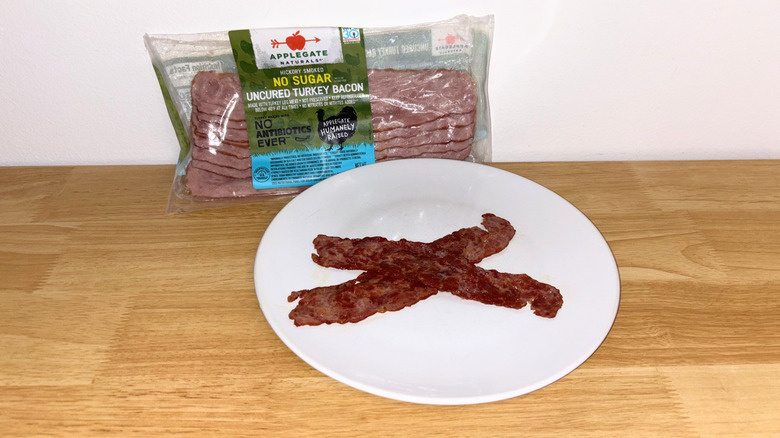 Two pieces of Applegate Naturals turkey bacon laid across each other on a white plate with the package behind it.