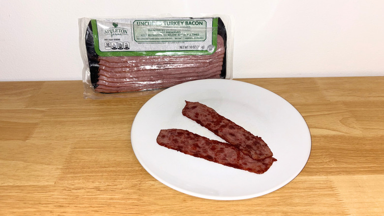 Two pieces of Appleton Farms turkey bacon laid across each other on a white plate with the package behind it.