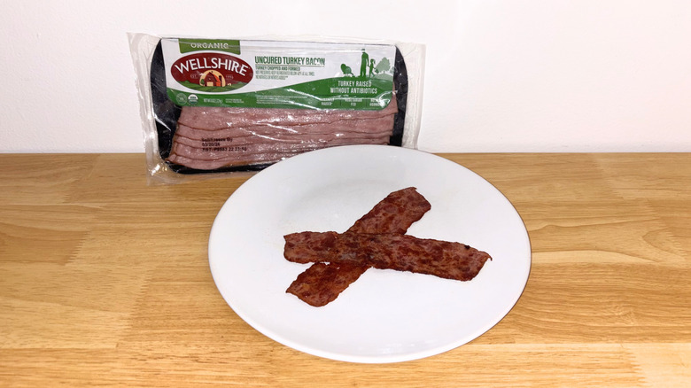 Two pieces of Wellshire turkey bacon laid across each other on a white plate with the package behind it.