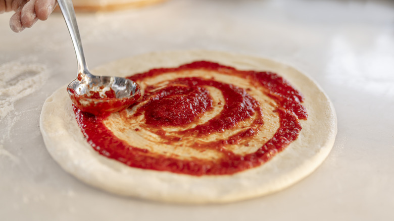 A hand ladeling tomato sauce on raw pizza dough
