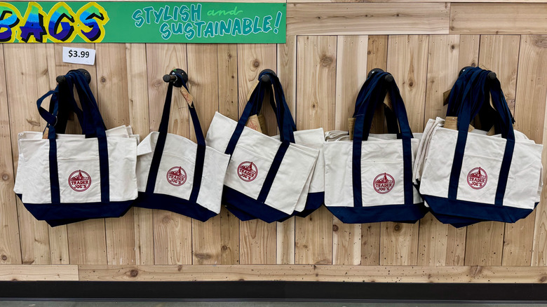 A line of reusable Trader Joe's tote bags hangs against a wooden wall with a sign that says "Bags, stylish and sustainable"