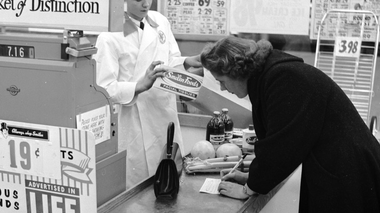 A black-and-white photo of a woman writing a check at a grocery store checkout in the 1950s