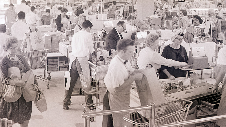 A black-and-white photo of a checkout section of grocery store in the 1960s with baggers loading paper bags into carts