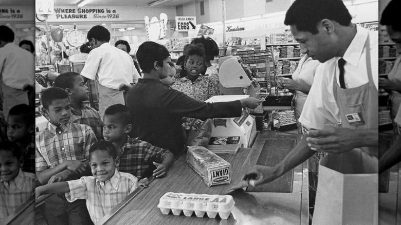Cashier placing groceries in a paper bag while children wait at a 1960s grocery store checkout lane
