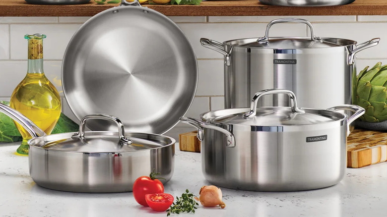 Tramontina stainless steel cookware on a kitchen counter surrounded by oil and various vegetables and herbs