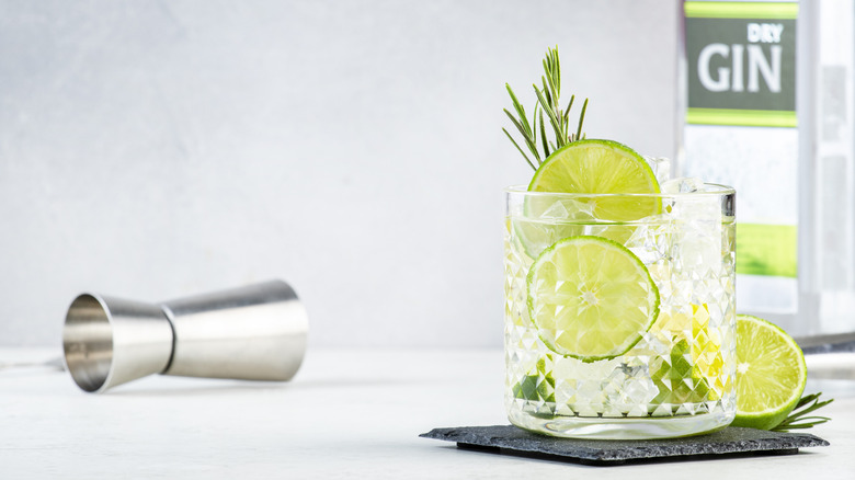 A gin cocktail with a lime wheels and a rosemary swig next to a jigger lying on its side