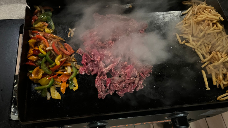 Peppers, beef, and fries cooking on an outdoor griddle