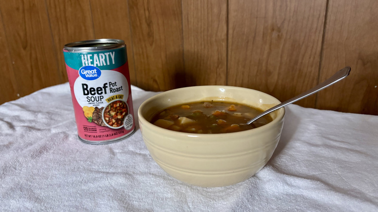 An empty can of Great Value Beef Pot Roast Soup behind a beige bowl filled with stew, with a spoon sticking out of it