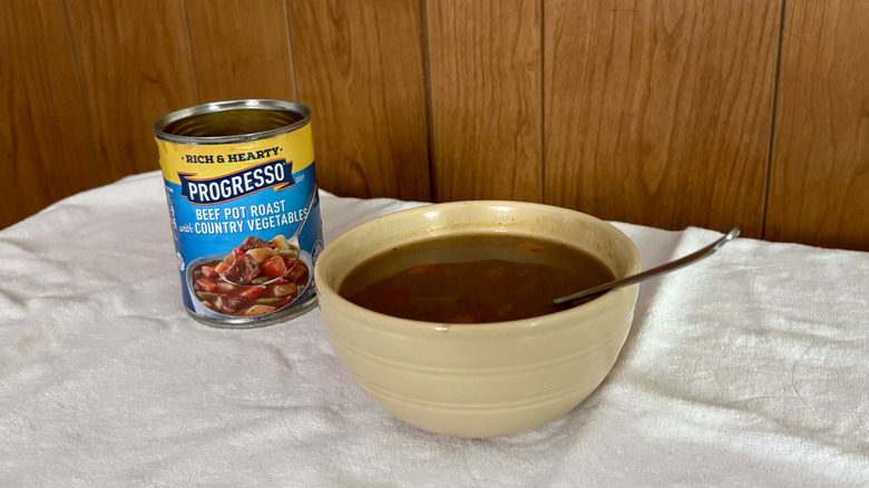 An empty can of Progresso Beef Pot Roast with Country Vegetables behind a beige bowl filled with stew, with a spoon sticking out of it