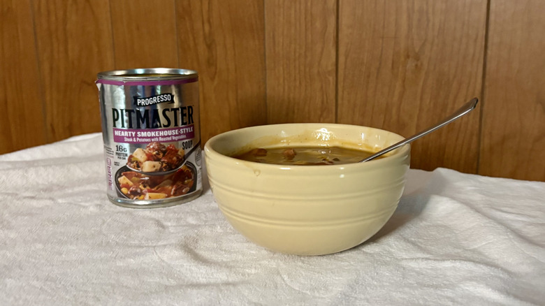 An empty can of Pitmaster Hearty Smokehouse-Style stew behind a beige bowl filled with stew, with a spoon sticking out of it