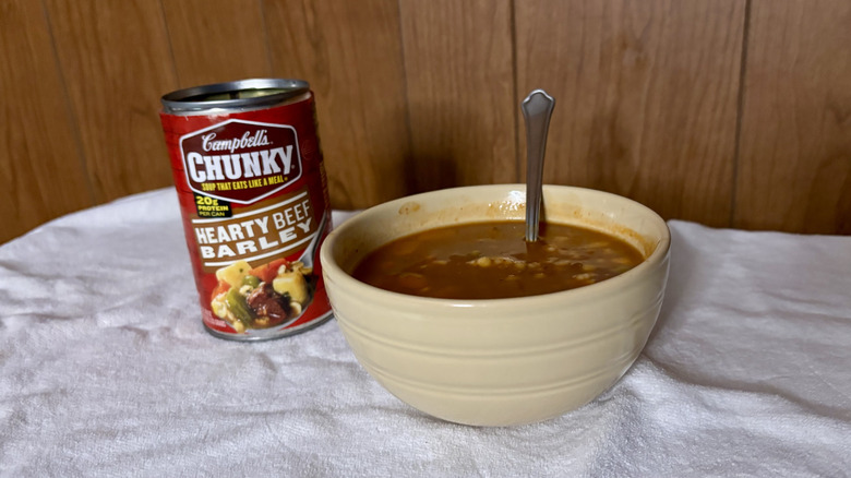 An empty can of Campbell's Chunky Hearty Beef Barley Stew behind a beige bowl filled with stew, with a spoon sticking out of it