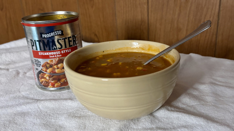 An empty can of Pitmaster Steakhouse-Style beef stew behind a beige bowl filled with stew, with a spoon sticking out of it