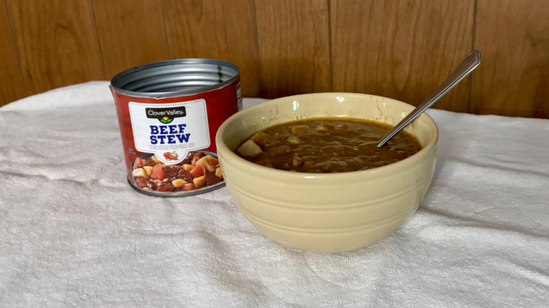 An empty can of Clover Valley Beef Stew behind a beige bowl filled with stew, with a spoon sticking out of it