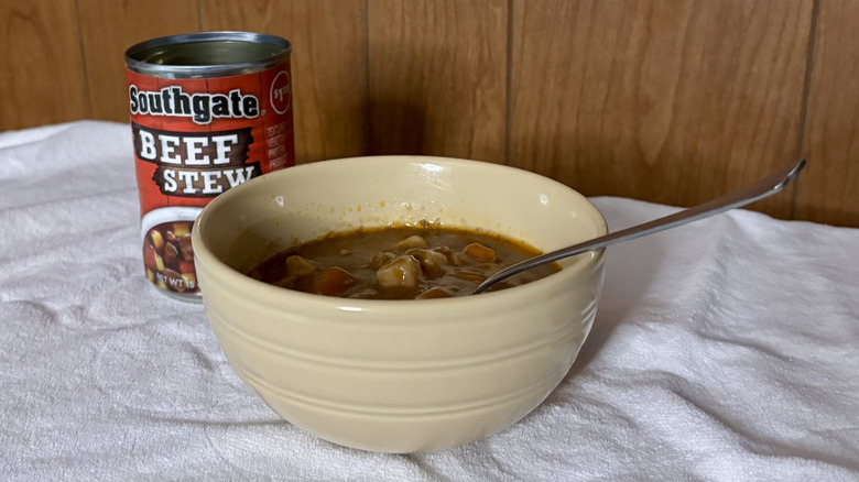 An empty can of Southgate Beef Stew behind a beige bowl filled with stew, with a spoon sticking out of it