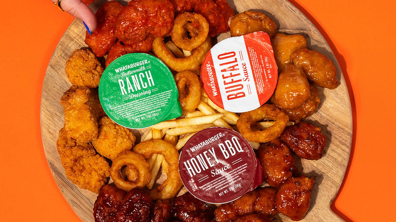 A wooden board full of Whataburger boneless wings, onion rings, fries, and sauce on an orange background