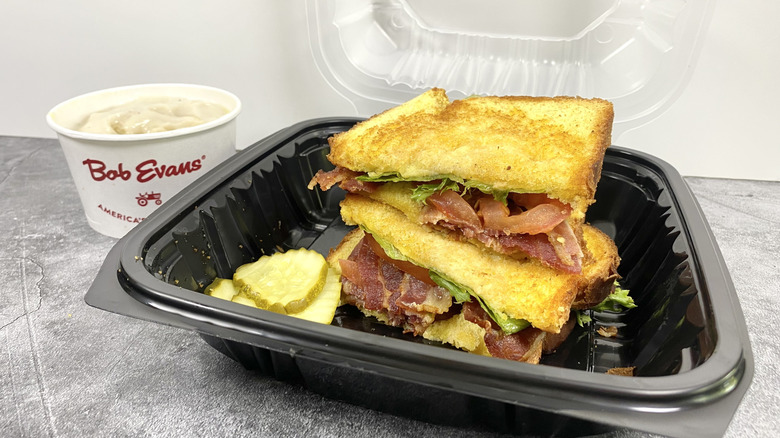 The Bob Evans All American BLT in its to-go container with pickles next to a side of mashed potatoes