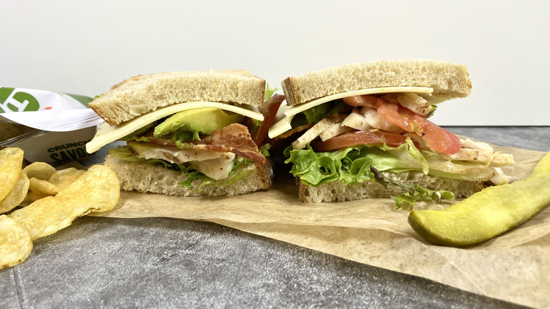 The Panera Bread Grilled Chicken & Avo BLT on top of its packaging next to chips and a pickle spear
