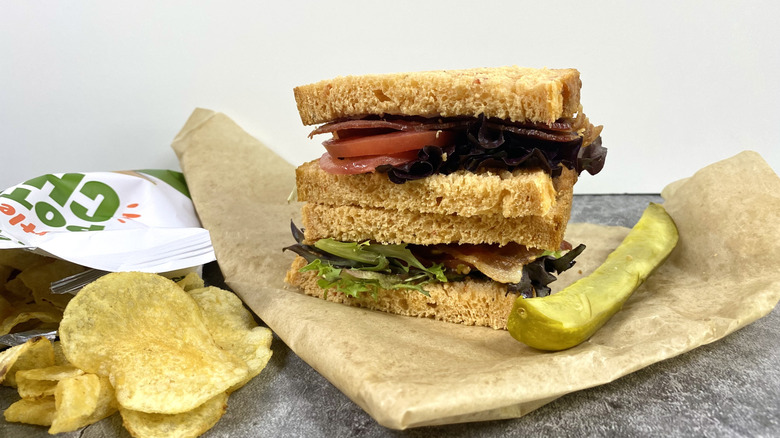 The Panera Bread Tomato Basil BLT on top of its paper packaging next to chips and a pickle spear