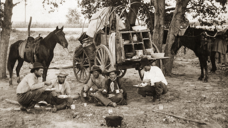 cowboys sitting and eating beside a chuck wagon