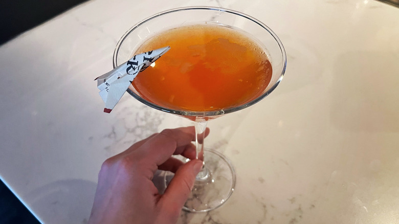 The Paper Plane from Bonefish Grill on a marble counter with a hand holding the stem and a tiny paper airplane stuck on the rim