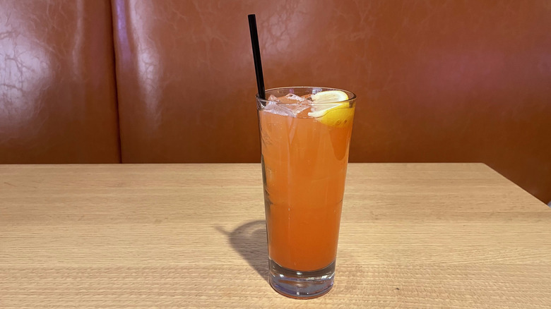 A grapefruit colored drink in a Collins glass with a lemon wedge sits on a wooden table.