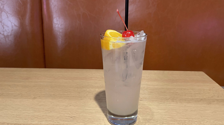 A collins glass filled with opaque liquid, a orange slice, and maraschino sits on a wooden table.