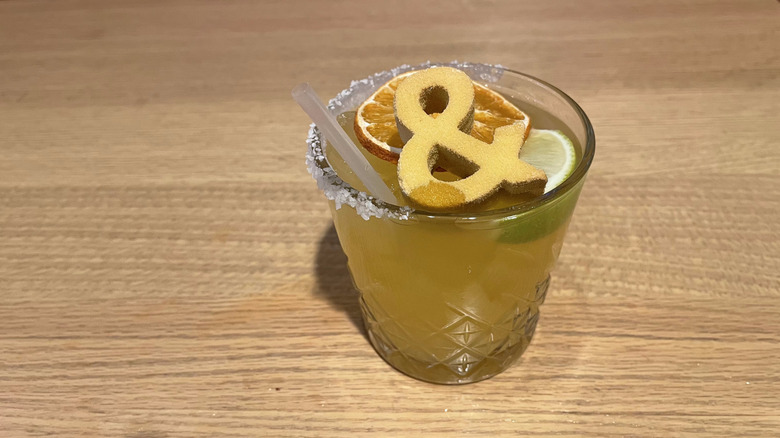 A margarita with a golden ampersand on top sits on a wooden table.