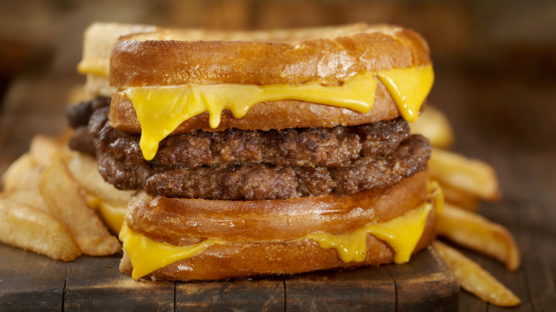 Two burger patties between two grilled cheese sandwiches with fries in background